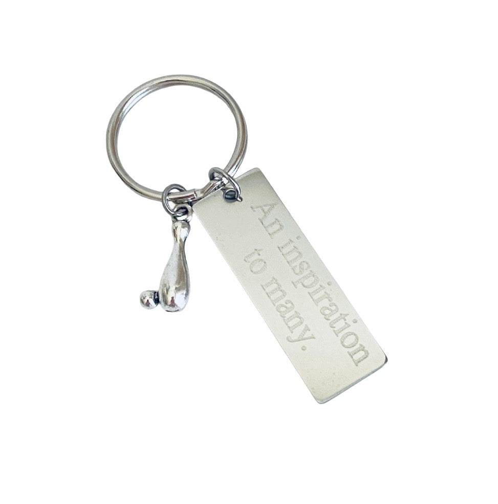 A silver metal keychain featuring a rectangular engraved tag that reads 'An inspiration to many.' and a small dangling bell, shown on a plain white background.
