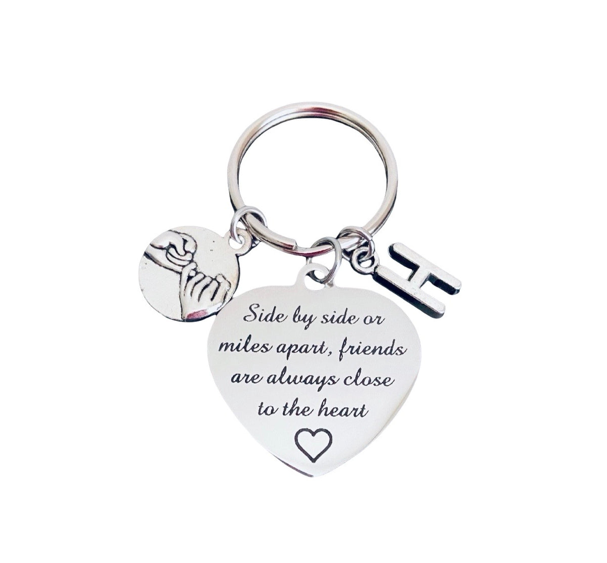 A silver metal keychain featuring a heart-shaped charm engraved with 'Side by side or miles apart, friends are always close to the heart' and a small circular charm showing two figures embracing, alongside a letter 'H' charm, on a plain white background.