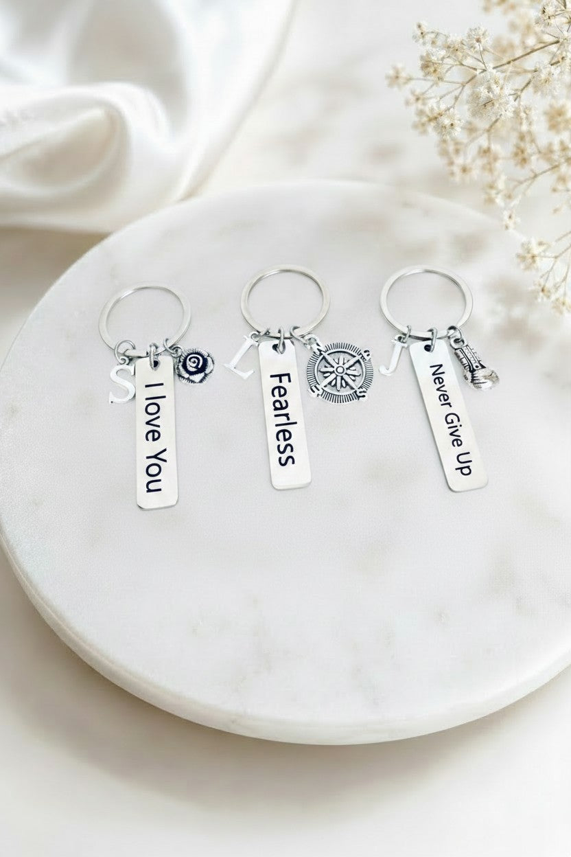 A pair of gold hoop earrings with white freshwater pearl beads and dangling gold initial letters, displayed on a round white marble tray with decorative dried flowers and white fabric.