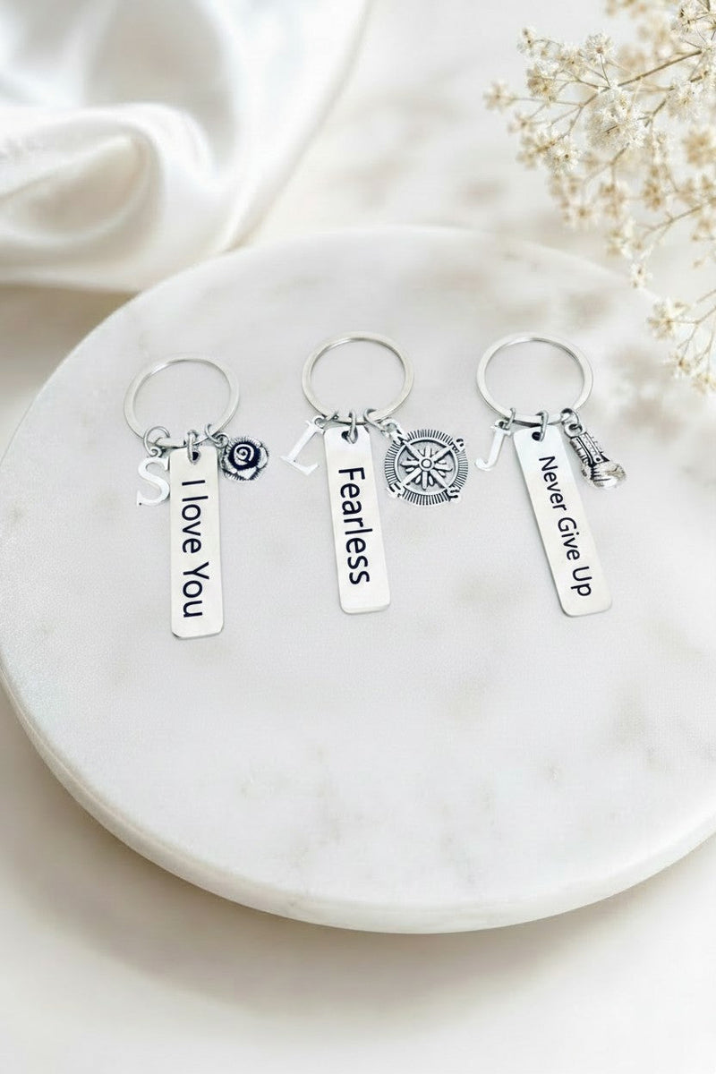 A pair of gold hoop earrings with white freshwater pearl beads and dangling gold initial letters, displayed on a round white marble tray with decorative dried flowers and white fabric.
