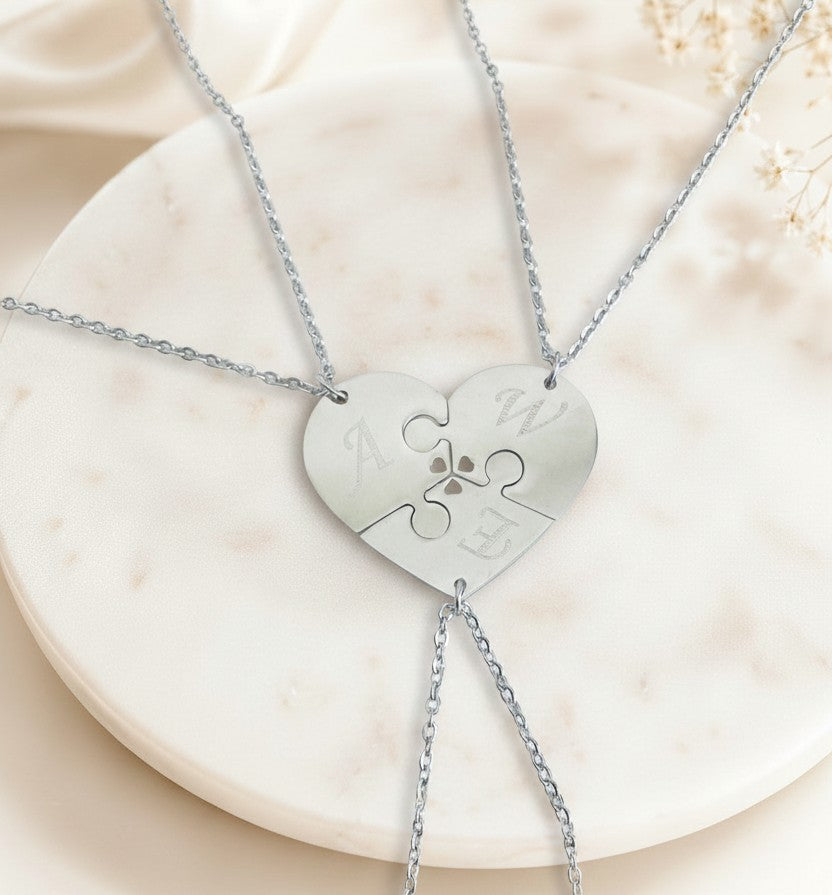 A three-piece silver puzzle heart necklace set displayed on a round white marble tray. The center of the puzzle pieces features a small engraved paw print, and the outer edges of the puzzle pieces are customized with silver initials (A, W, E).
