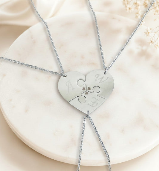 A three-piece silver puzzle heart necklace set displayed on a round white marble tray. The center of the puzzle pieces features a small engraved paw print, and the outer edges of the puzzle pieces are customized with silver initials (A, W, E).