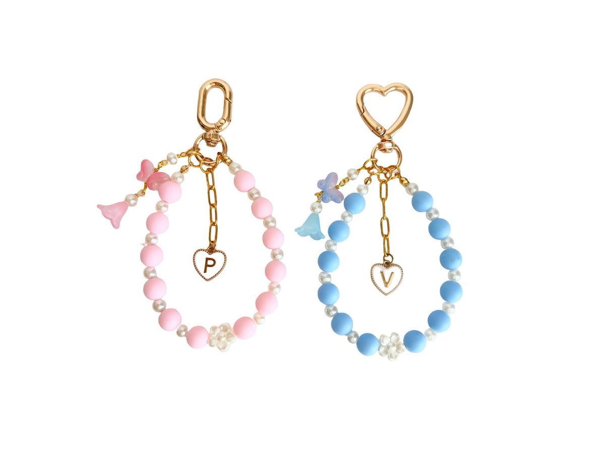 Two gold-tone beaded hoop earrings displayed side-by-side on a white background. The left earring features light pink beads, small white pearl-like beads, a pink bow charm, and a dangling heart charm with the letter 'P'.