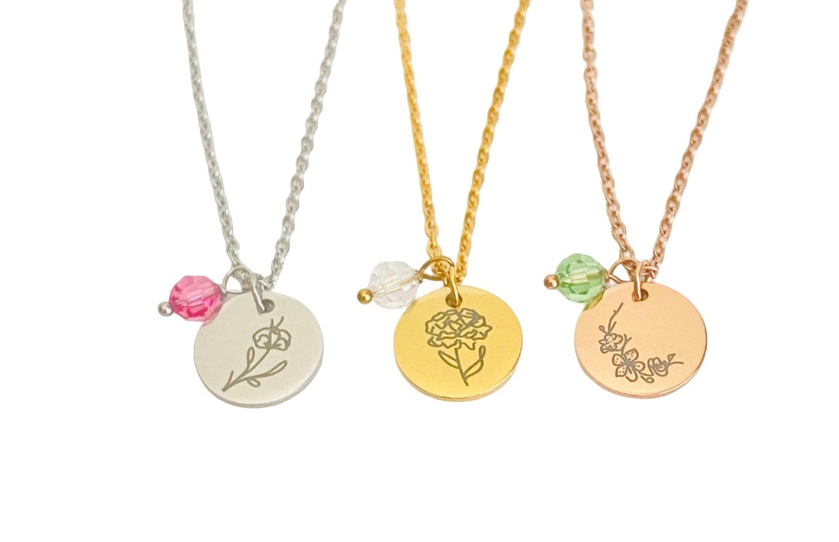 Three floral pendant necklaces on delicate chains, shown side by side on a white background. The necklaces feature round engraved flower pendants in silver, gold, and rose gold tones, each accented with a small faceted bead in pink, clear, and light green respectively.