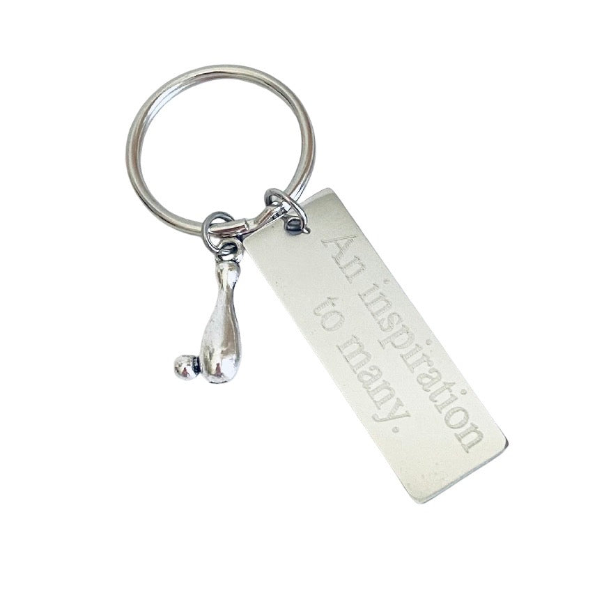 A silver metal keychain featuring a rectangular engraved tag that reads 'An inspiration to many.' and a small teardrop-shaped charm, shown on a plain white background.