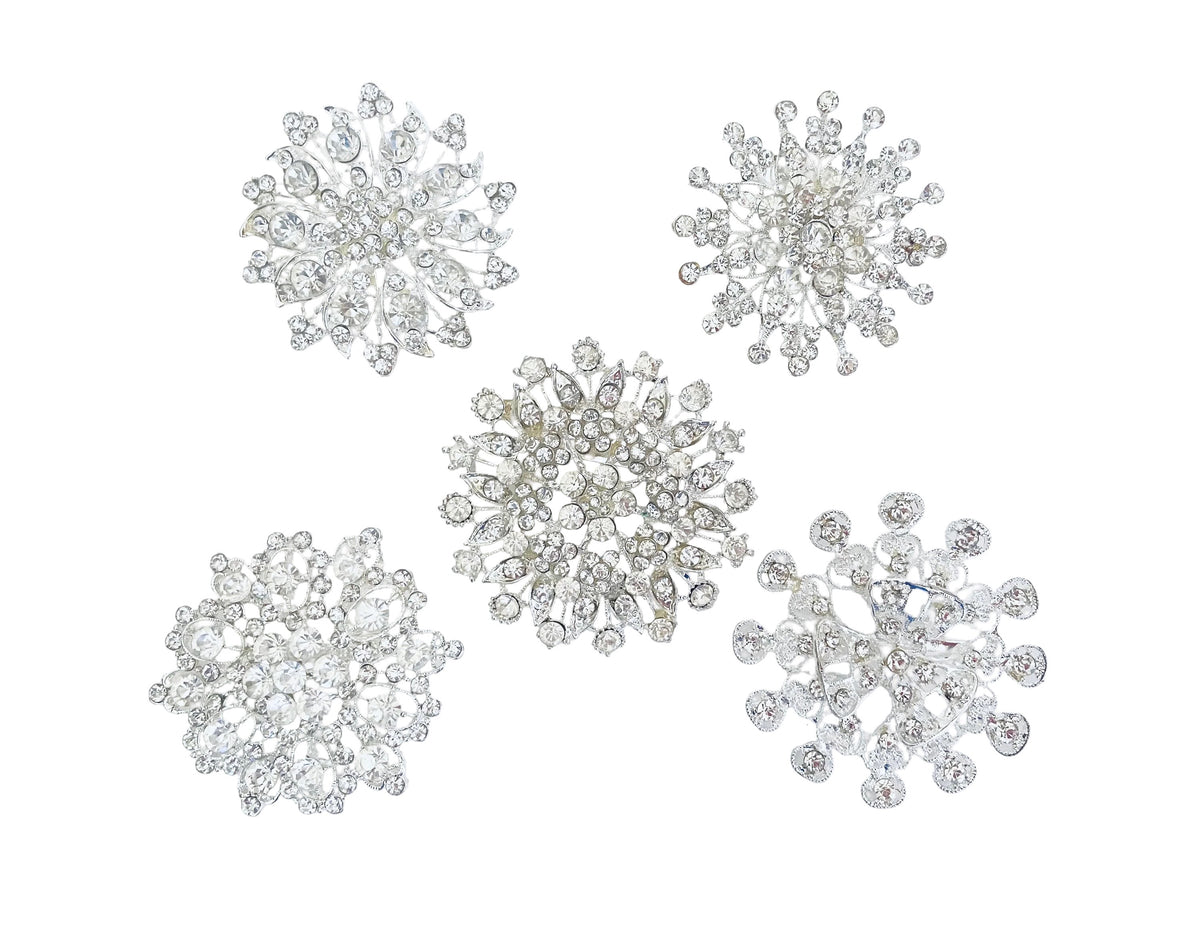 Five silver snowflake-shaped brooches embellished with clear crystals, arranged on a plain white background.