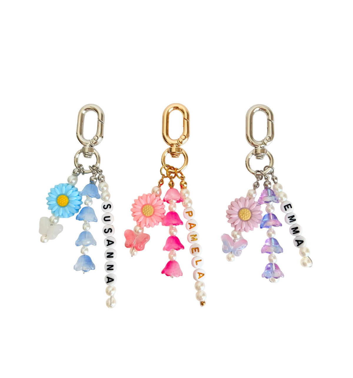 Three floral dangle earrings with silver and gold-tone clasp tops, decorated with pastel blue, pink, purple, and white flowers, beads, pearls, butterflies, and vertical text reading SUSANNA, PAMELA, and EMMA on a white background.