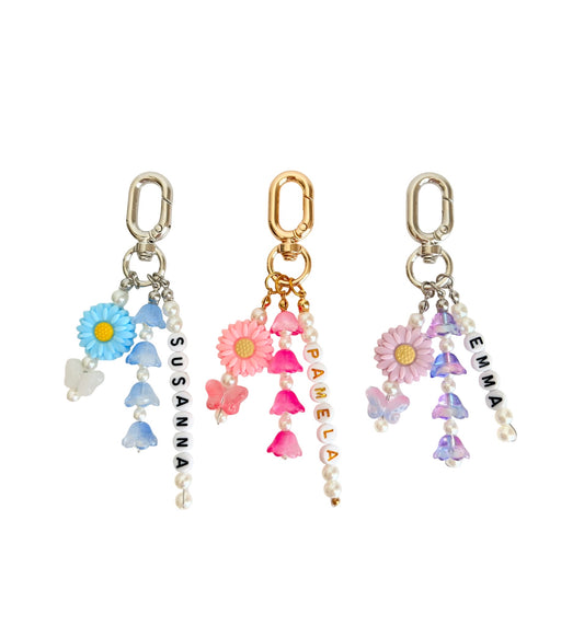 Three floral dangle earrings with silver and gold-tone clasp tops, decorated with pastel blue, pink, purple, and white flowers, beads, pearls, butterflies, and vertical text reading SUSANNA, PAMELA, and EMMA on a white background.