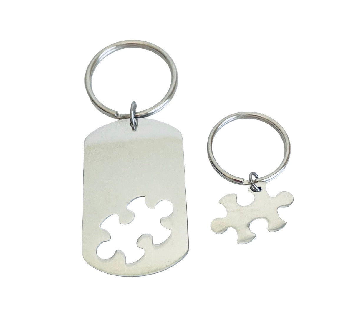 Two silver metal keychains on a plain white background: one larger dog tag-style keychain with a puzzle-piece cutout and one smaller matching puzzle charm keychain.