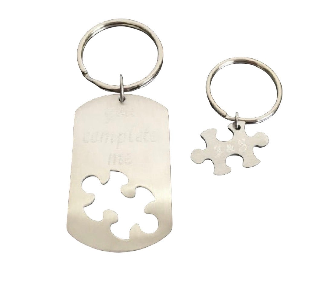 Two silver metal keychains on a plain white background: one larger dog tag-shaped keychain with a puzzle cutout and faint engraved text, and one smaller puzzle-piece keychain.
