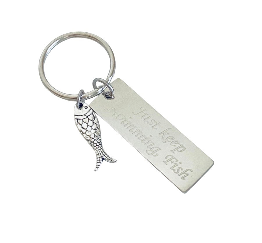 A silver-toned metal keychain featuring a round split ring, a rectangular engraved nameplate that reads 'Just Keep Swimming, Fish', and a small fish charm, shown on a plain white background.