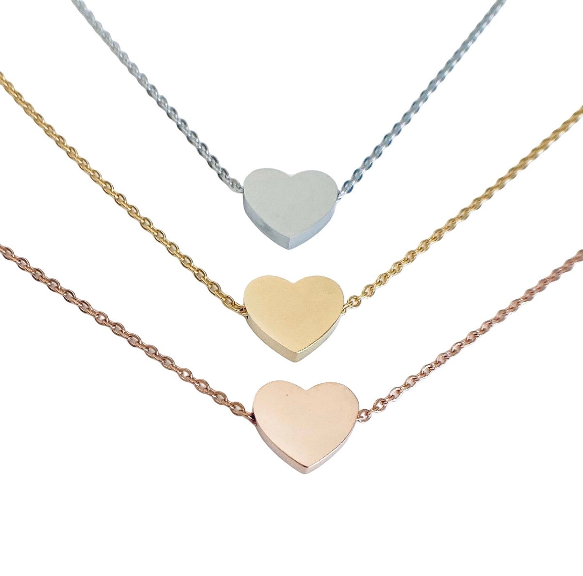 Three heart-shaped pendant necklaces on delicate chains, shown against a plain white background. The top necklace is silver, the middle is yellow gold, and the bottom is rose gold.
