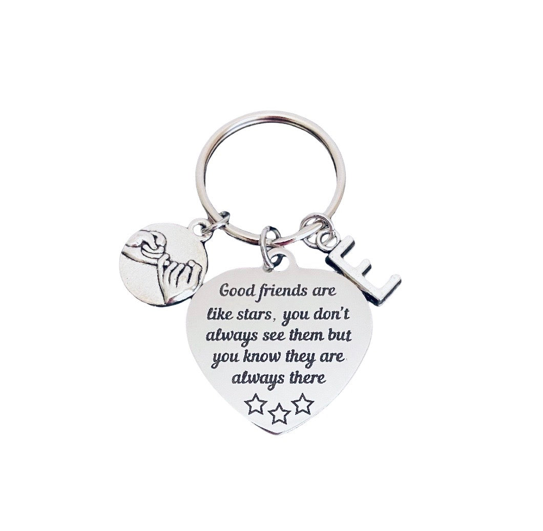 A silver metal keychain with three charms: a heart-shaped pendant engraved with 'Good friends are like stars, you don't always see them but you know they are always there' and three stars, a small round charm showing two hands holding, and a small letter 'E' charm, all attached to a split key ring on a white background.