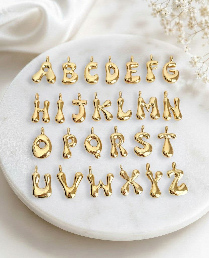 Gold metallic alphabet letter charms arranged in three rows on a round white marble tray, with soft white fabric and dried flowers in the background.