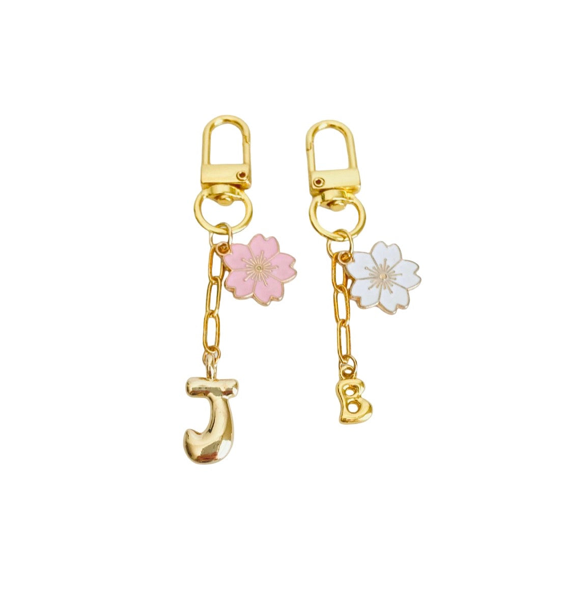 A pair of gold-tone drop earrings with spring rings, featuring a pink cherry blossom charm, a white cherry blossom charm, and a dangling gold initial 'J' charm, shown on a plain white background.