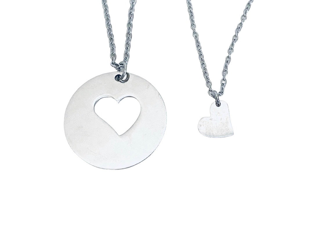 Two silver heart-themed necklaces on a white background: one with a large round disc pendant featuring a heart-shaped cutout, and one with a small textured heart pendant on a chain.