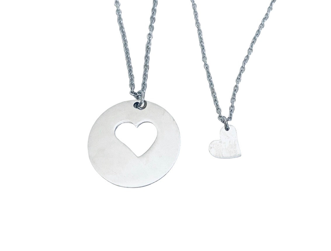 A silver necklace featuring a large circular disc pendant with a heart-shaped cutout, shown next to a smaller textured heart pendant on a matching chain, against a plain white background.