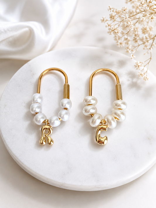 A pair of gold hoop earrings with white freshwater pearl beads and dangling gold initial letters, displayed on a round white marble tray with decorative dried flowers and white fabric.