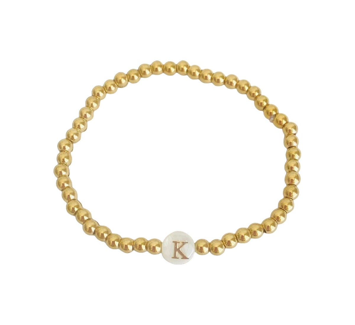 A gold beaded stretch bracelet featuring a small white oval charm engraved with the letter 'K', displayed on a clean white background.