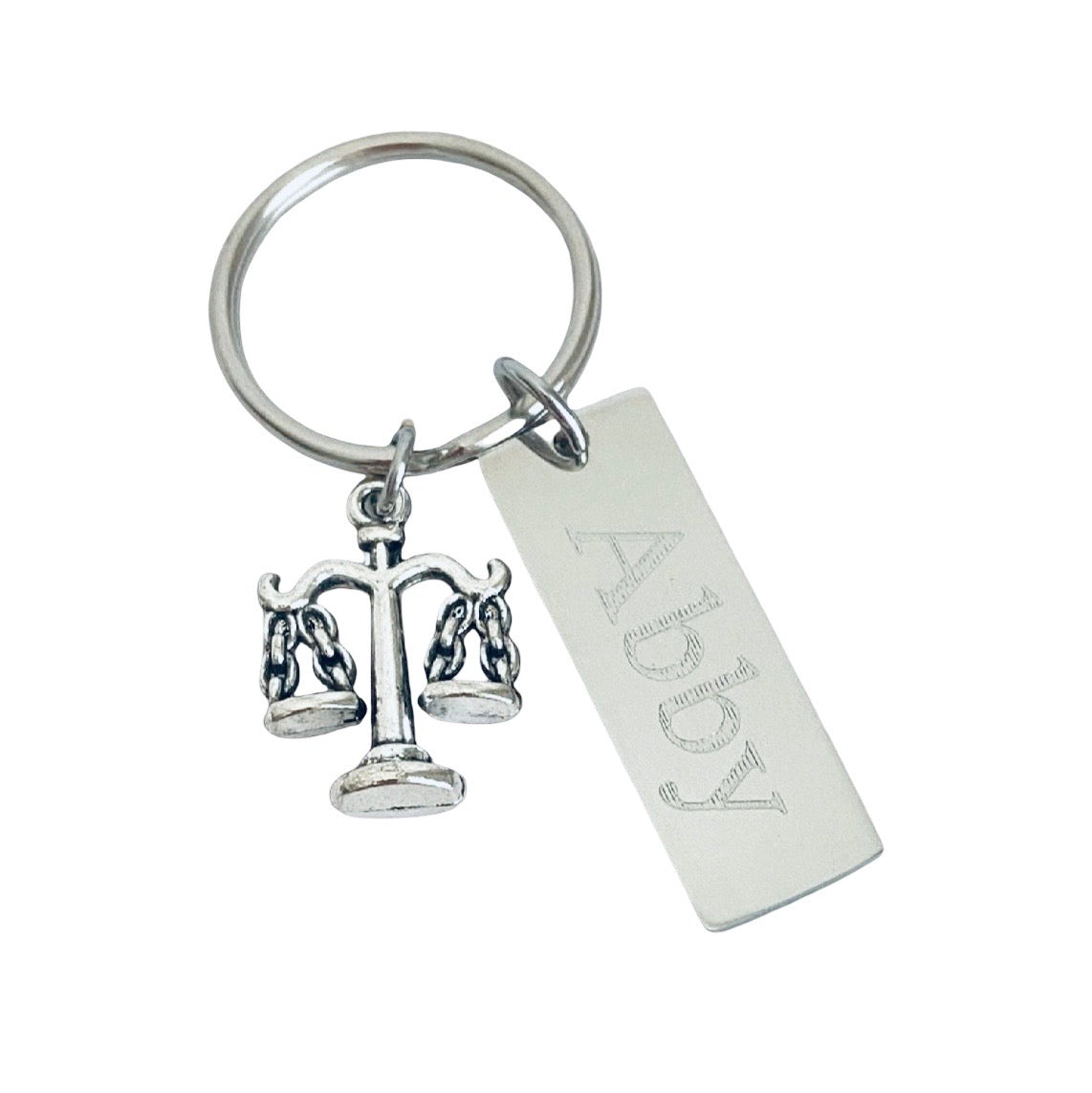 A silver-toned metal keychain featuring a small balance scale charm and a rectangular engraved tag, shown on a plain white background.