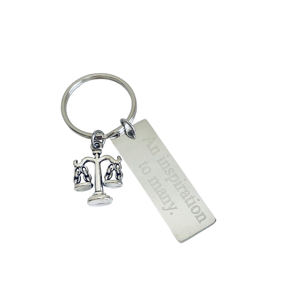 A silver-toned metal keychain featuring a small scales of justice charm and a rectangular engraved tag that reads 'An inspiration to many.' on a plain white background.