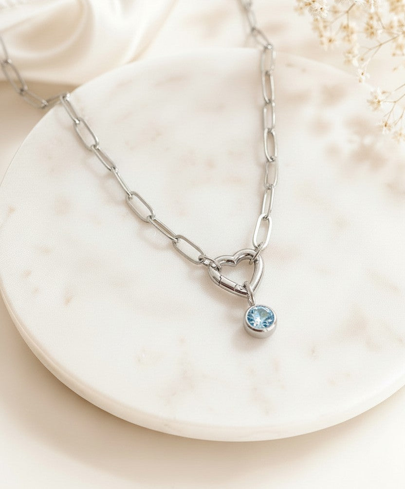 A silver paperclip chain necklace with a heart-shaped pendant and a small round blue gemstone, displayed on a round white marble tray.