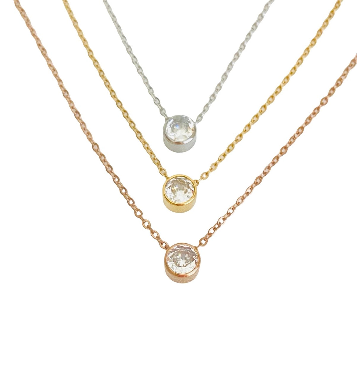 Three delicate necklace chains in rose gold, yellow gold, and silver, each featuring a bezel-set round clear gemstone pendant, arranged diagonally on a white background.