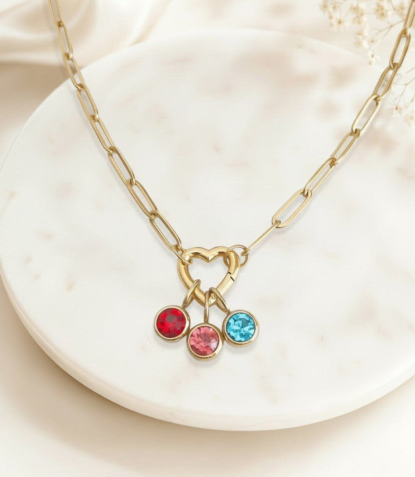 Gold chain necklace with a heart-shaped clasp and three dangling round gemstone charms in red, pink, and blue, displayed on a white marble plate.