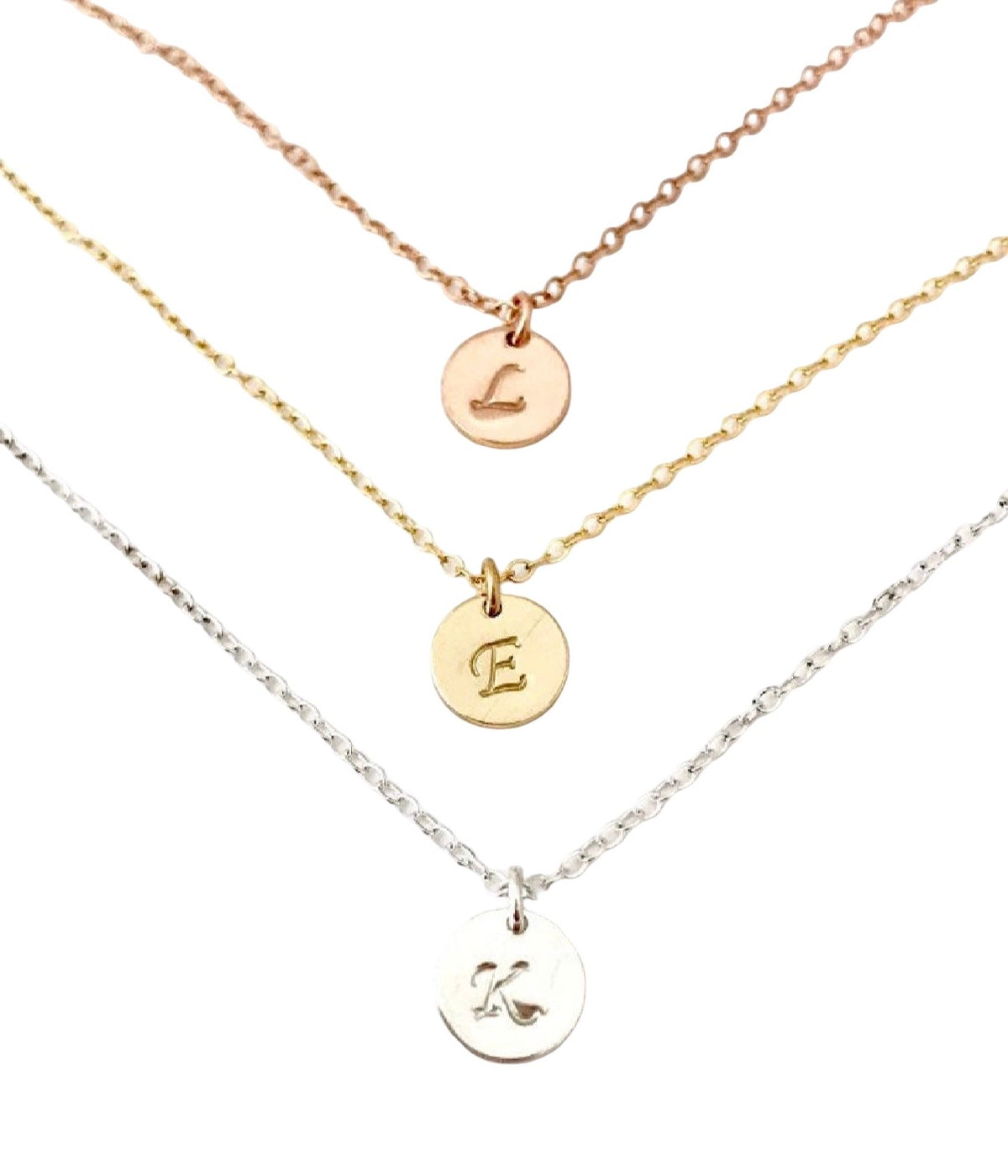 Three delicate chain necklaces with round initial pendants are arranged diagonally on a white background. The top necklace is rose gold, the middle is gold, and the bottom is silver, each with a small engraved letter.